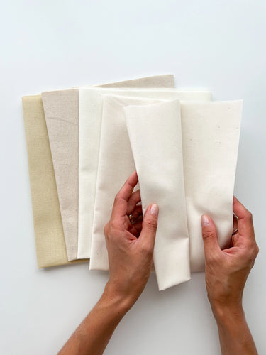 A hand holds four fat quarters of cotton and linen-cotton blend fabrics in neutral colors.