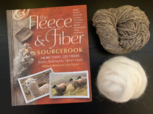 Load image into Gallery viewer, The Fleece and Fiber Sourcebook, a large hardcover book with photos of alpaca, sheep and fiber on its cover, lies next to a white ball of wool roving and a gray ball of yarn with white speckles.
