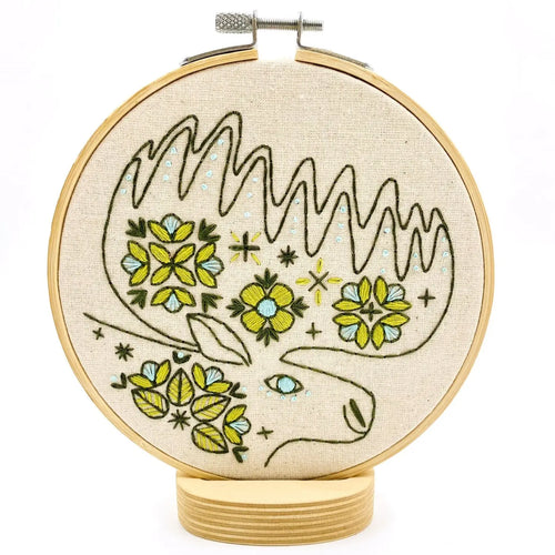 Embroidery hoop holds unbleached fabric stitched with a folk-style moose design, in greens and blues.