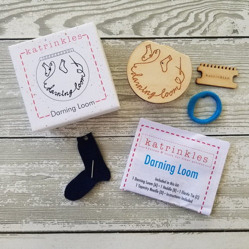 A Katrinkles small darning loom box and contents - the rounded wood darning loom, wood heddle teeth, needle and felt sock holder, fabric elastic band, and instructions.