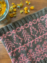 Load image into Gallery viewer, A colorwork knit cowl lies on a table next to a glass canning jar of dried yellow calendula blossoms; it is knit in a main color of golden brownish green, with a contrast color of pink depicting trees, flowers and vines.
