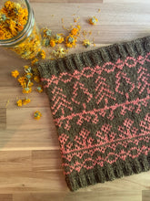 Load image into Gallery viewer, A colorwork knit cowl lies on a table next to a glass canning jar of dried yellow calendula blossoms; it is knit in a main color of golden brownish green, with a contrast color of pink depicting trees, flowers and vines.
