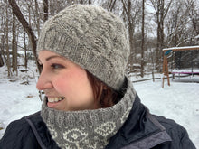 Load image into Gallery viewer, A woman stands in the snow wearing a gray wool hat, knit with cables of alternating thickness and a ribbed brim.
