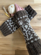 Load image into Gallery viewer, A hand holds a white mini pumpkin and wears a brown knit armwarmer almost to the elbow, with ribbing at either end and a white colorwork motif of vines and flowers. The other armwarmer and several stems of dried wheat lie beneath the hand on the table. 
