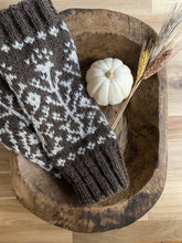 Load image into Gallery viewer, A pair of brown knit armwarmers, with ribbing at either end and a white colorwork motif of vines and flowers, lie in a carved wood bowl beside a white mini pumpkin and several stems of dried wheat.
