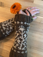 Load image into Gallery viewer, A pair of hands hold an orange calendula blossom and wear a pair of brown knit armwarmers almost to the elbow, with ribbing at either end and a white colorwork motif of vines and flowers.
