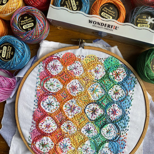 The Yo Yo Rainbow Sampler - a pre-printed sampler in many stripes of colors, with circles that can be filled in and stitched over - in an embroidery hoop surrounded by embroidery floss.