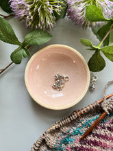 Load image into Gallery viewer, Pink ceramic bowl holding silver acorn stitch markers.
