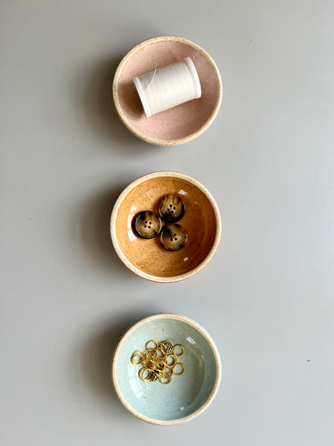 Three ceramic notions bows in pale pink, terra cotta, and sky blue, holding spool of thread, buttons and gold stitch markers.