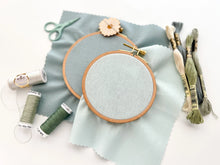 Load image into Gallery viewer, Two wood embroidery hoops hold linen-cotton fabrics in medium and light blue, surrounded by embroidery threads of blue and green.
