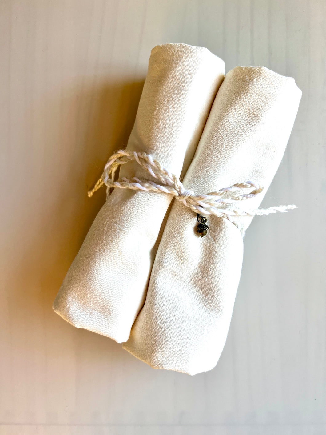 Two natural flour sack towels rolled up and tied together with decorative twine and a small brass acorn charm.