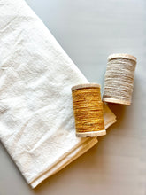 Load image into Gallery viewer, A folded natural flour sack towel lies beside yellow and white spools of wool thread.
