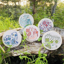 Load image into Gallery viewer, Five embroidery hoops hold unbleached fabric stitched with folk patterns of moose, polar bears, caribou, fox, and wolves, resting on a fallen log in the woods.
