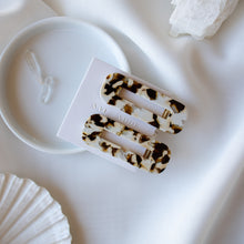 Load image into Gallery viewer, A pair of tortoise style hair clips in cream and dark brown lay on a white table cloth and saucer.
