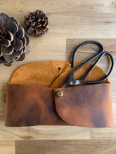 Load image into Gallery viewer, A rectangular leather pouch in lighter and darker patina leather with two curved sides and a rounded flap, held together with a brass clasp. A tapestry needle and steel scissors peek out the top. Pinecones lie nearby.
