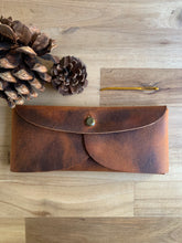 Load image into Gallery viewer, A rectangular leather pouch in lighter and darker patina leather with two curved sides and a rounded flap, held together with a brass clasp. Pinecones lie nearby.
