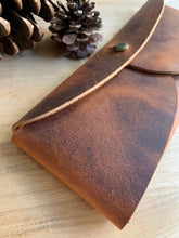 Load image into Gallery viewer, A rectangular leather pouch in lighter and darker patina leather with two curved sides and a rounded flap, held together with a brass clasp. Pinecones lie nearby.
