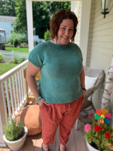 Load image into Gallery viewer, A woman with brown curly hair stands on a summer porch wearing an aqua knit tee and reddish linen pants.
