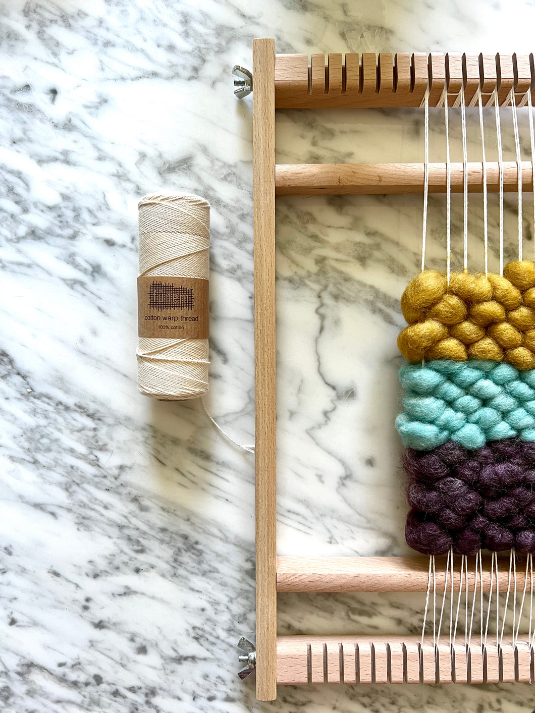 A roll of white cotton warp thread lies on a marble tabletop next to a small wood tapestry handheld loom, warped in the same cotton thread, with wefts of yellow, aqua and purple core rug yarn.