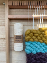 Load image into Gallery viewer, A roll of white cotton warp thread lies on a wood tabletop next to a small wood tapestry handheld loom, warped in the same cotton thread, with wefts of yellow, aqua and purple core rug yarn.
