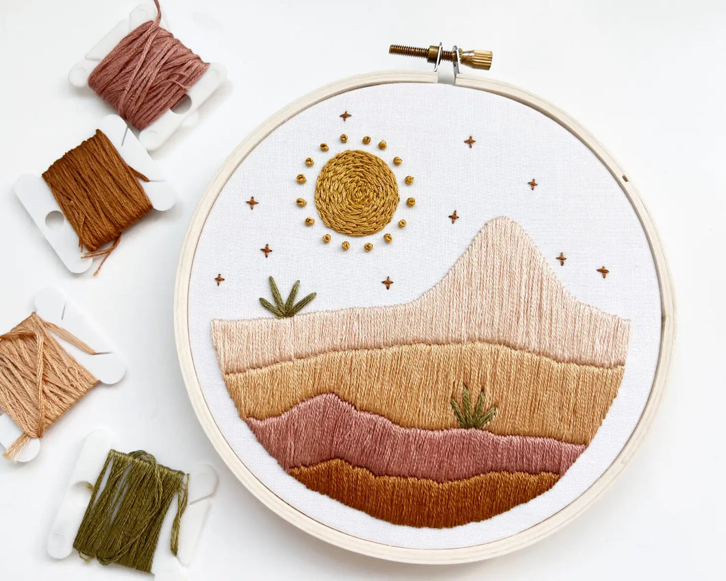 An embroidery hoop holds an image of desert plain and mountain in shades of orange and pink, with a sun and plants. 