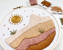 Load image into Gallery viewer, An embroidery hoop holds an image of desert plain and mountain in shades of orange and pink, with a sun and plants. 
