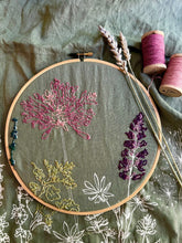 Load image into Gallery viewer, A 9&quot; wood embroidery hoop holds stitched flowers in pink, green and purple on a green linen tea towel, with two spools of pink thread lying nearby.
