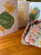 Load image into Gallery viewer, A set of 3 cards are standing next to a gray colorwork knit in progress. The cards have 3 distinct linocut designs in green, yellow and purple, depicting a flower and skein; a skein with a loose strand prickling with little leaves, or vegetable matter; and a basket made of two leaves holding a pair of knitting needles and 3 skeins.
