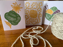 Load image into Gallery viewer, Three cards with distinct linocut designs in green, yellow and purple stand on a table next to an oatmeal yarn skein that is caked. The linocut images are a green skein with a yellow flower behind it; a yellow skein with a strand unraveling, prickling with small leaves, or vegetable matter; and a green basket formed by two leaves, holding a pair of purple knitting needles and three skeins of yarn in green, yellow and purple.
