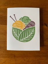 Load image into Gallery viewer, A card with a linocut design depicting a green basket made of two leaves, holding a pair of purple knitting needles and three skeins of yarn in yellow, purple and green.
