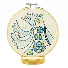 Load image into Gallery viewer, A wood embroidery hoop holds unbleached fabric stitched with a folk-style caribou in blues and yellows.
