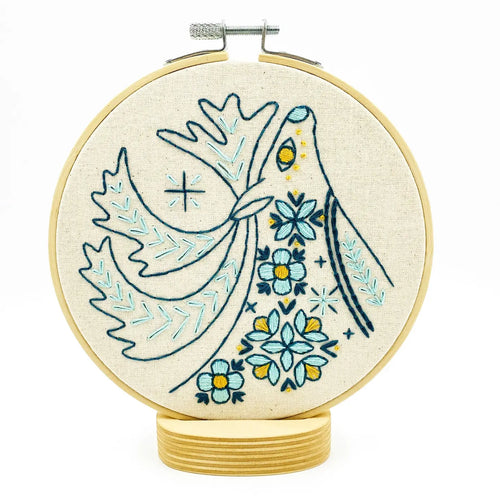 A wood embroidery hoop holds unbleached fabric stitched with a folk-style caribou in blues and yellows.