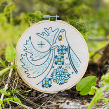 Load image into Gallery viewer, A wood embroidery hoop holds unbleached fabric stitched with a folk-style caribou in blues and yellows.
