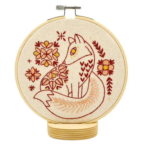 An embroidery hoop holds unbleached fabric stitched with a fox surrounded by flowers, in red, pink and yellow threads.