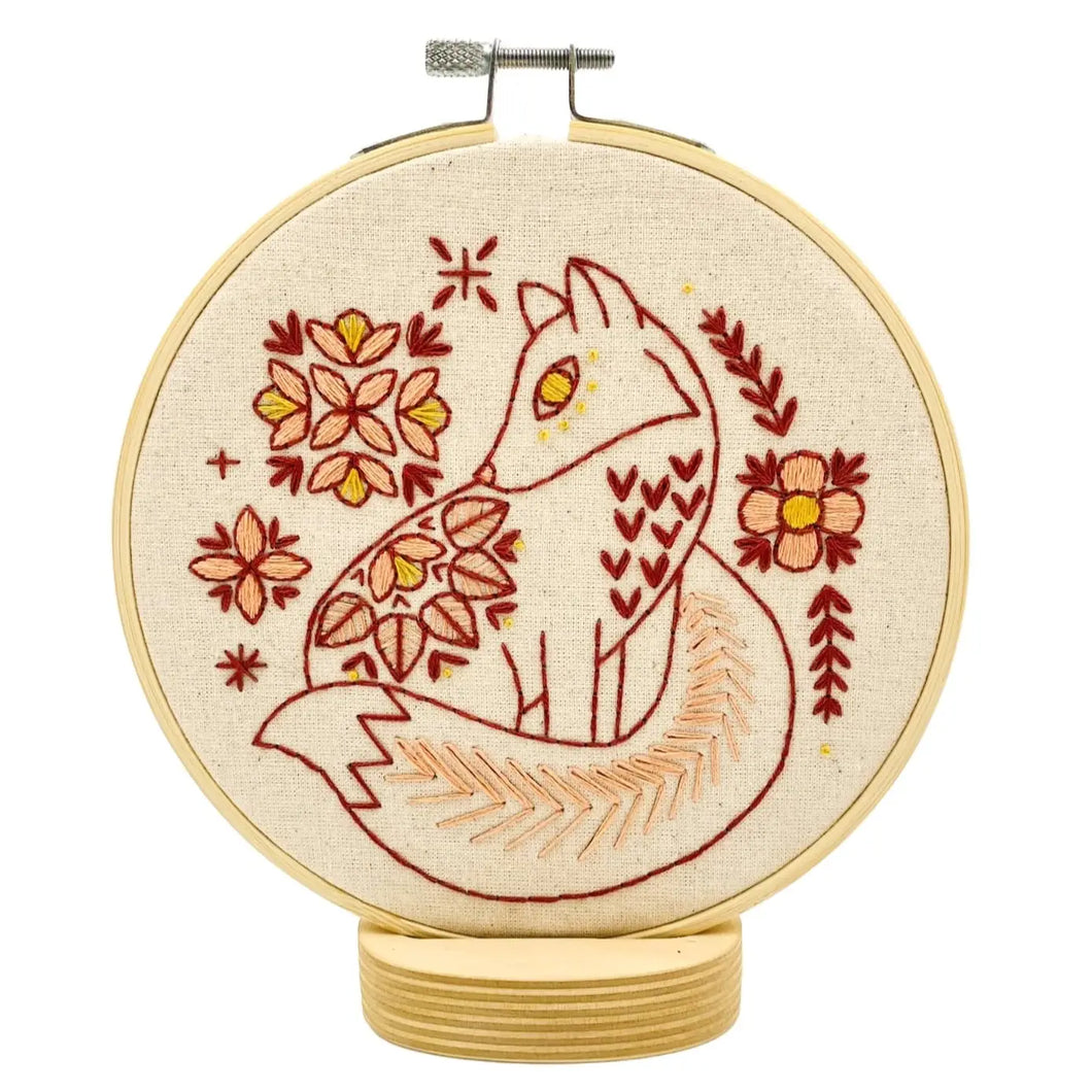 An embroidery hoop holds unbleached fabric stitched with a fox surrounded by flowers, in red, pink and yellow threads.