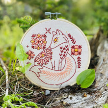 Load image into Gallery viewer, An embroidery hoop holds unbleached fabric stitched with a fox surrounded by flowers, in red, pink and yellow threads.
