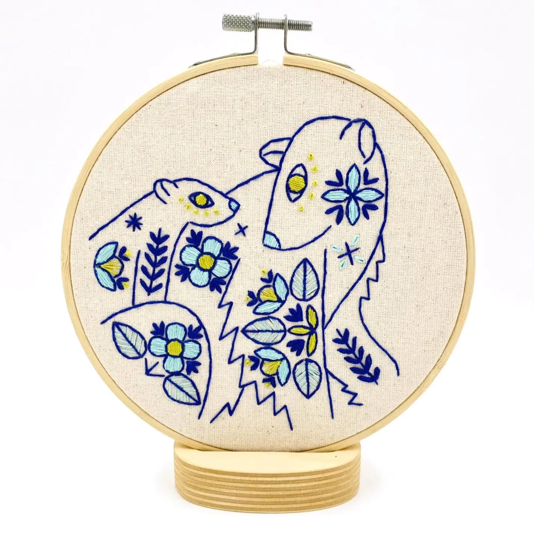 A wood embroidery hoop holds unbleached fabric stitched with a pattern of mother and baby polar bear in blues and yellow, accented with folk-style floral motifs.