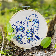 Load image into Gallery viewer, A wood embroidery hoop holds unbleached fabric stitched with a pattern of mother and baby polar bear in blues and yellow, accented with folk-style floral motifs.
