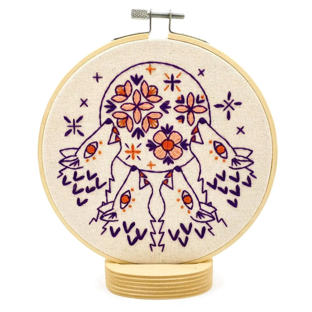 A wood embroidery hoop holds unbleached fabric stitched with a pattern of four wolves howling at a moon decorated with flowers, in colors of purple, red and pink.