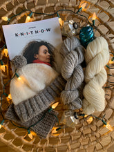 Load image into Gallery viewer, A skein of silvery gray and white yarn, a pair of gray mitts and gray and white hat knit from the same yarn, and the book Knit How lay in a large, flat, circular basket surrounded by white twinkle lights and several mercury glass ornaments.
