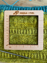 Load image into Gallery viewer, A 4&quot; square gauge swatch ruler with centimeter and inches measurements, and the Needle + Purl logo at the top, lays atop a yellow and blue knit piece.
