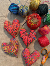 Load image into Gallery viewer, Four red fabric hearts embroidered with multi colored threads. Scissors, a pincushion and spools of thread lie nearby.
