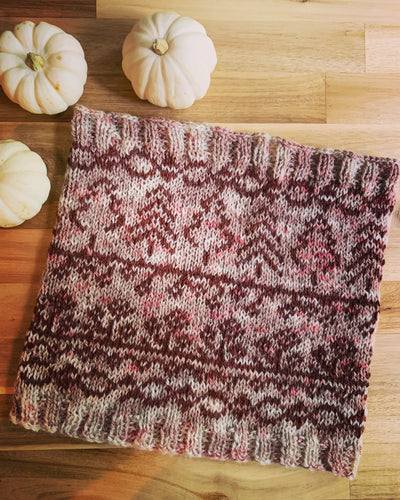 A colorwork knit cowl lies on a table next to 3 white mini pumpkins; it is knit in a main color of varying mauves and pinks with a contrast color of dark mauve depicting trees, flowers and vines.