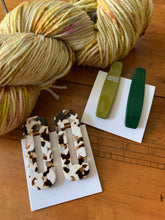 Load image into Gallery viewer, A pair of tortoise style hair clips in brown and cream, and another pair of clips in shades of green, lie next to a speckled skein of yellow yarn.
