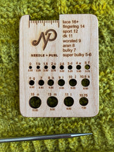 Load image into Gallery viewer, A needle gauge tool with the Needle + Purl logo includes holes for measuring knitting needles, and a guide for measuring yarn gauge. It lays atop a yellow knitted piece.
