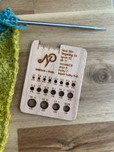 Load image into Gallery viewer, A needle gauge tool with the Needle + Purl logo includes holes for measuring knitting needles, and a guide for measuring yarn gauge. It lays next to a yellow knitted piece and metal knitting needle.
