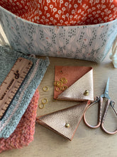 Load image into Gallery viewer, Two rose gold leather triangular stitch marker pouches lie on a table next to knit swatches, rose gold scissors, and a fabric Praktical project bag. One pouch is open and gold stitch markers spill out onto the table.
