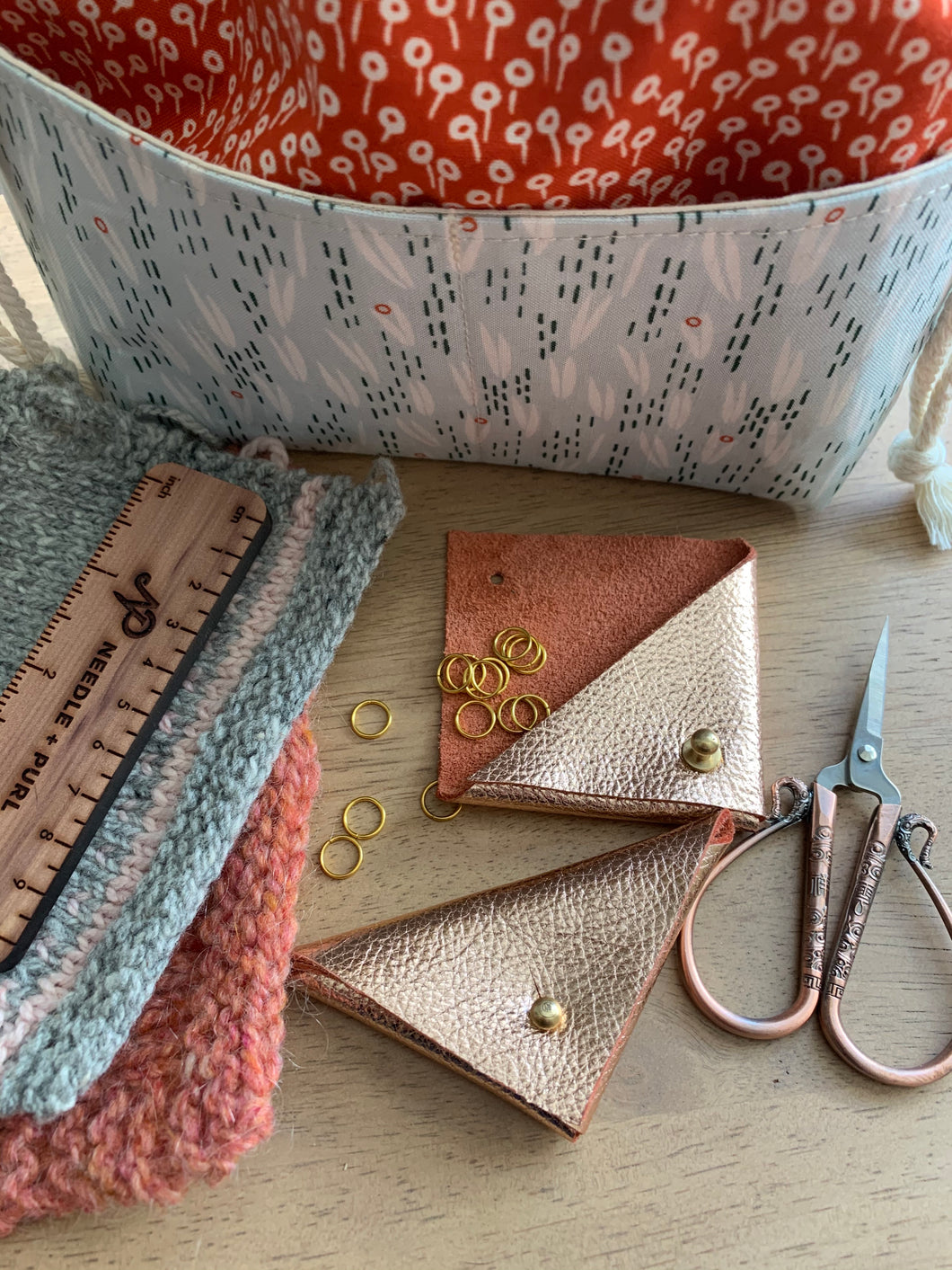 Two rose gold leather triangular stitch marker pouches lie on a table next to knit swatches, rose gold scissors, and a fabric Praktical project bag. One pouch is open and gold stitch markers spill out onto the table.
