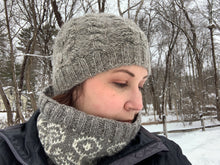 Load image into Gallery viewer, A woman stands in the snow wearing a gray wool hat, knit with cables of alternating thickness and a ribbed brim.

