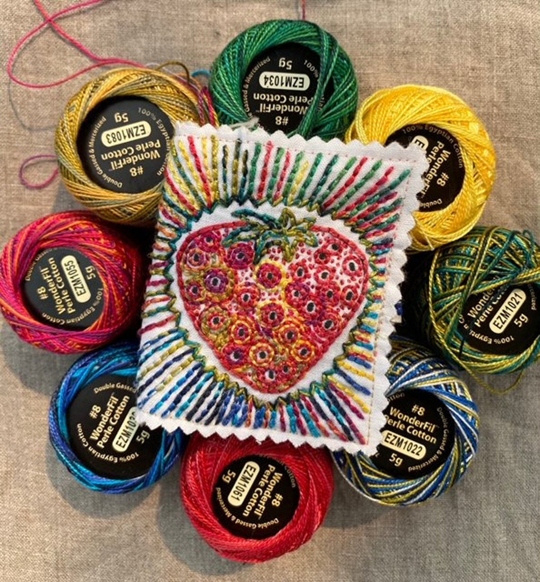 A small needlebook stitched with a strawberry and rainbow design, surrounded by spools of perle cotton thread.